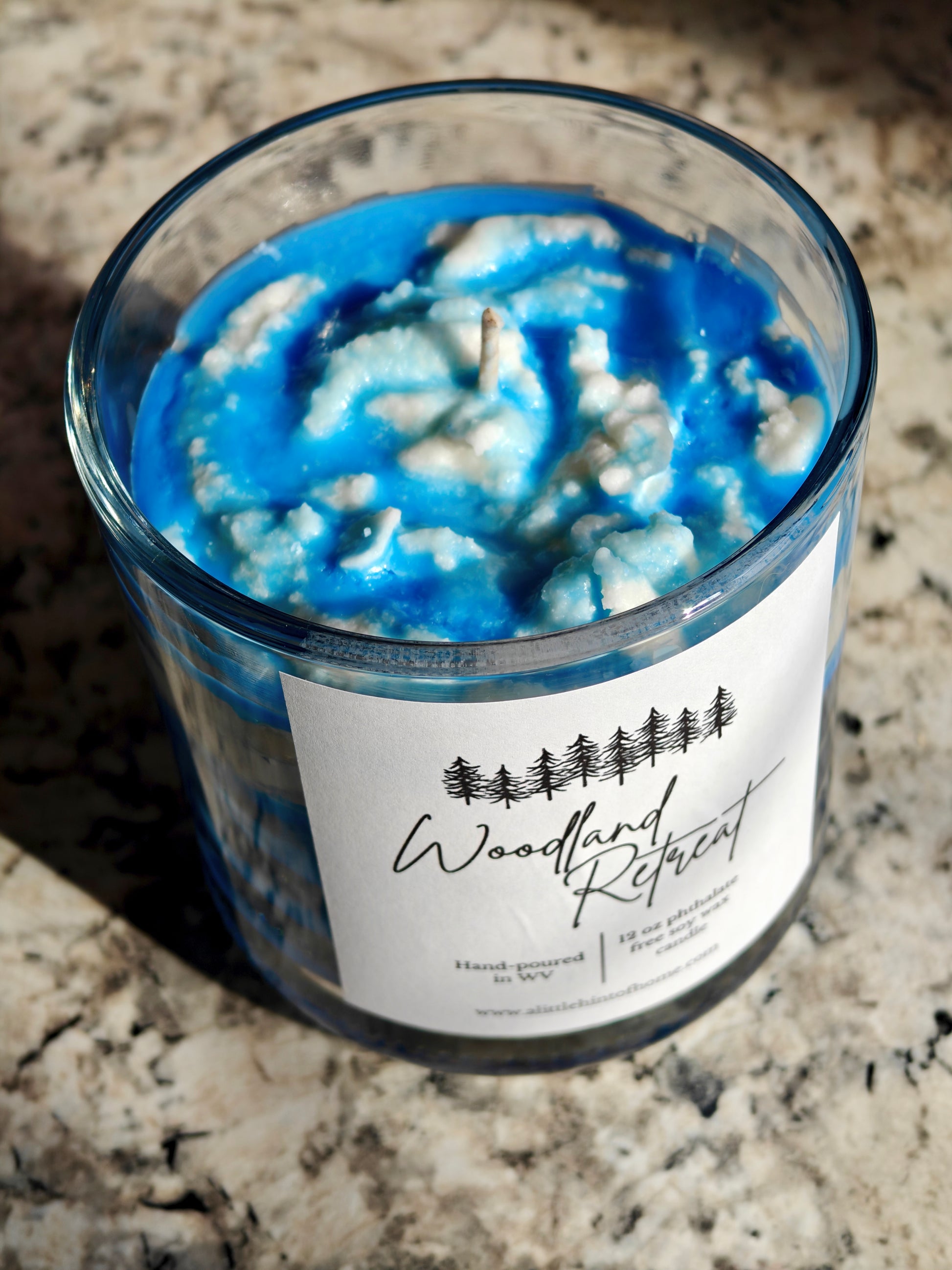 Candle with blue and white wax in a glass jar on a granite surface, labeled 'Woodland Retreat'.