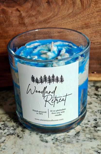 Candle with blue and white design in a glass jar on a marble surface, labeled 'Woodland Retreat'