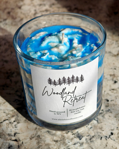 Candle with blue and white design in a glass jar on a marble surface, labeled 'Woodland Retreat'.